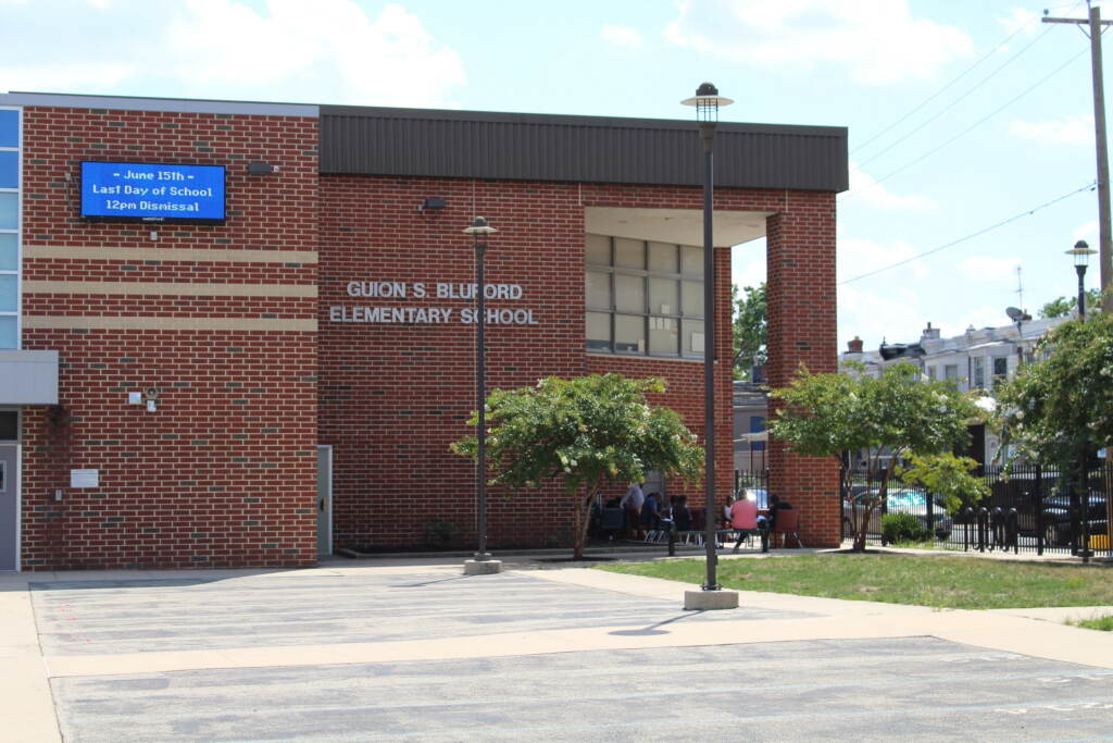 A brick building which reads Guion S. Bluford Elementary School is visible from the outside, with houses, trees and grass nearby visible to the right.