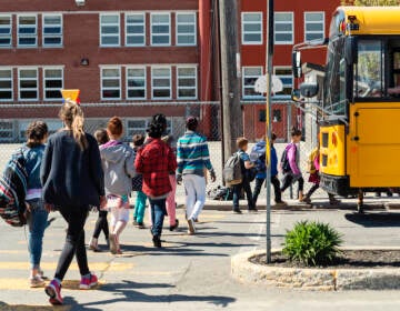 Students walk by a yellow school bus towards a red-brick school building.
