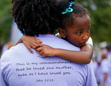 A little girl looks over the shoulder of an adult wearing a shirt which has a Bible verse written on the back.