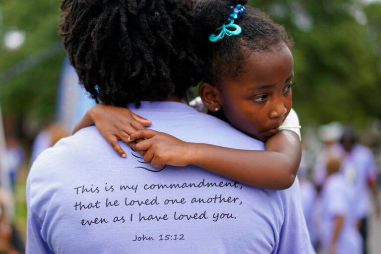A little girl looks over the shoulder of an adult wearing a shirt which has a Bible verse written on the back.