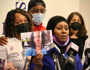 A woman holds up a picture of a woman as she speaks into several microphones. Three other people with somber faces are visible in the background.