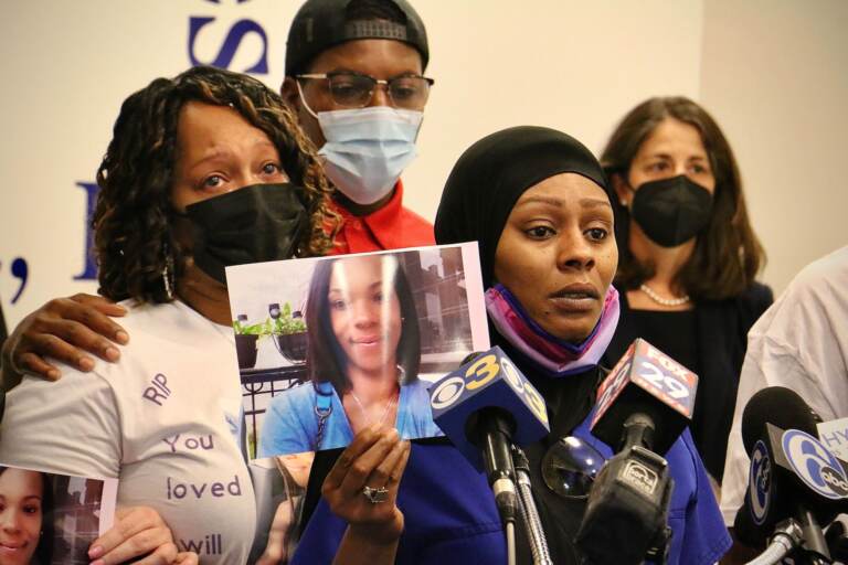 A woman holds up a picture of a woman as she speaks into several microphones. Three other people with somber faces are visible in the background.