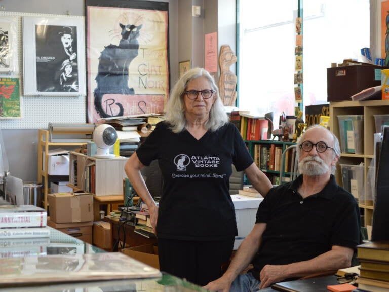 independent-bookstores-banned-books Two people, one sitting, one standing, look towards the camera, with bookshelves full of books visible behind them.