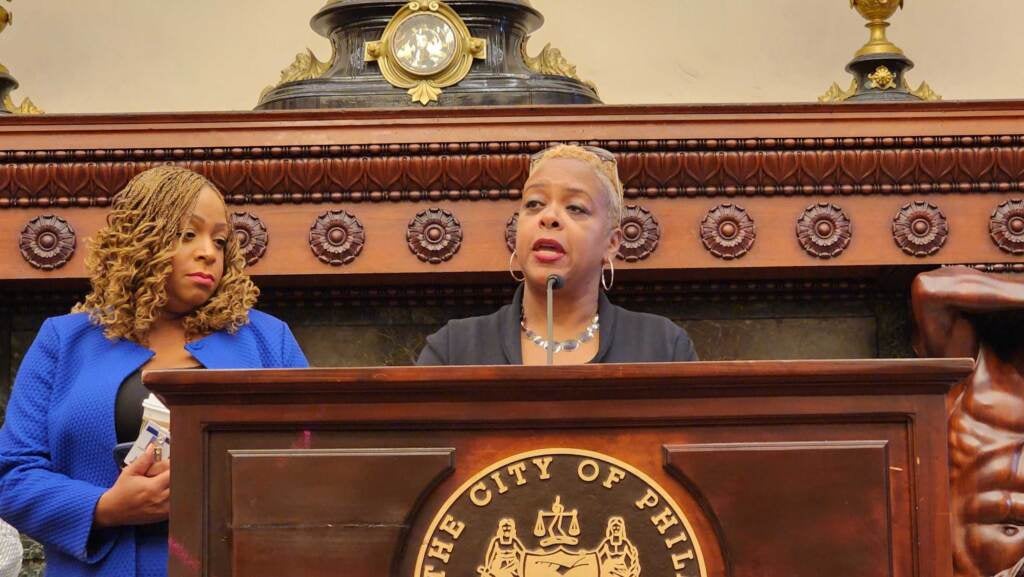 Cindy Bass speaks at a podium as Jamie Gauthier looks on.