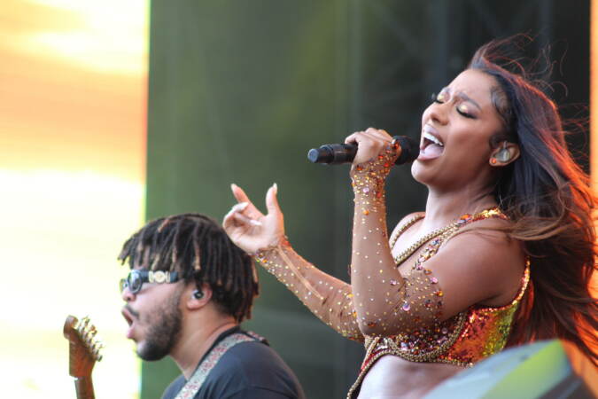 A singer sings into a microphone with her hair flowing behind her. A guitarist is visible in the background.