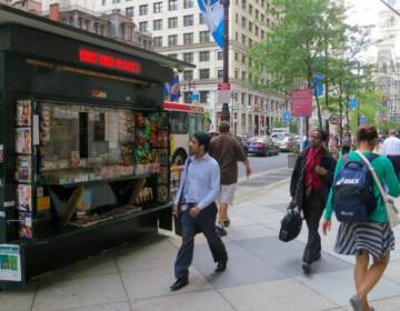 People walk by a newsstand on the sidewalk.