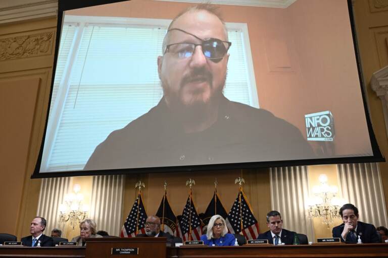 A person appears on a screen above the Jan. 6 commission members who are seated at a table.