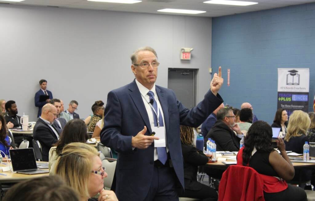 A man stands in the middle of a room full of people seated, gesturing as he speaks.