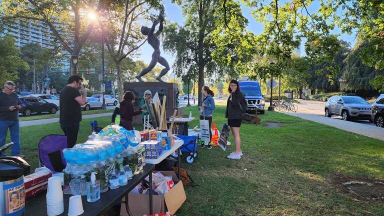 People gather around a table with water bottles and snacks. A sculpture, trees, and street are visible in the background.