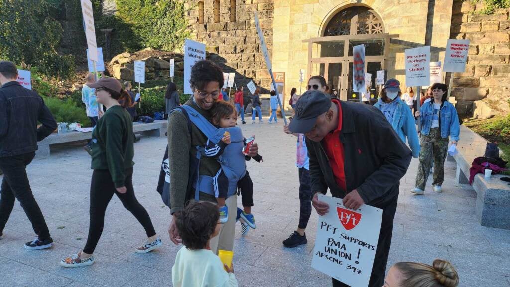 A man with two young kids speaks to another man as people holding signs walk in a circle in the background.