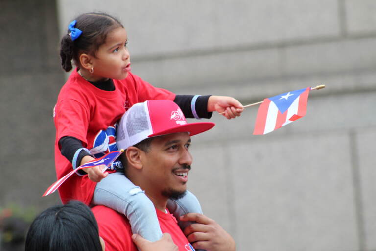 A little girl sits on her dad's shoulders, waving Puerto Rican flags in both hands.