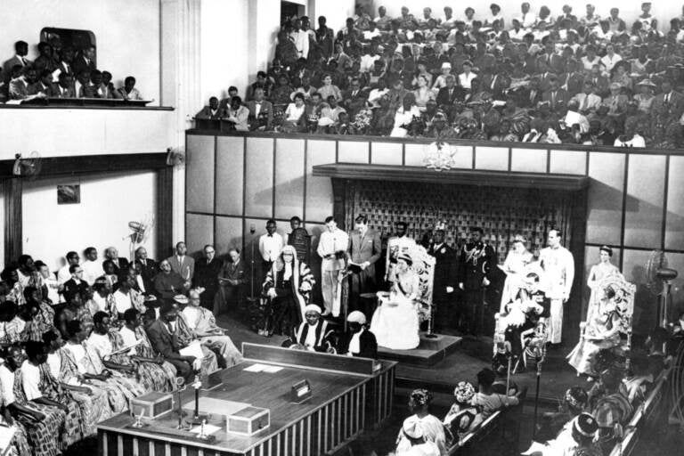 A black and white photo shows the Queen in a large room filled with people.