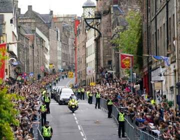 A hearse makes its way through the street in Scotland, which is lined with people on either side.