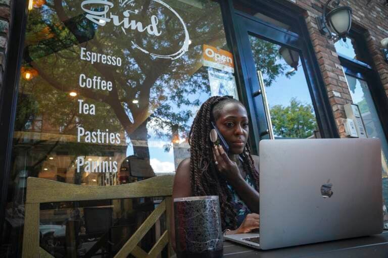 A woman holds a phone up to her ear while looking at a laptop screen outside of a storefront.