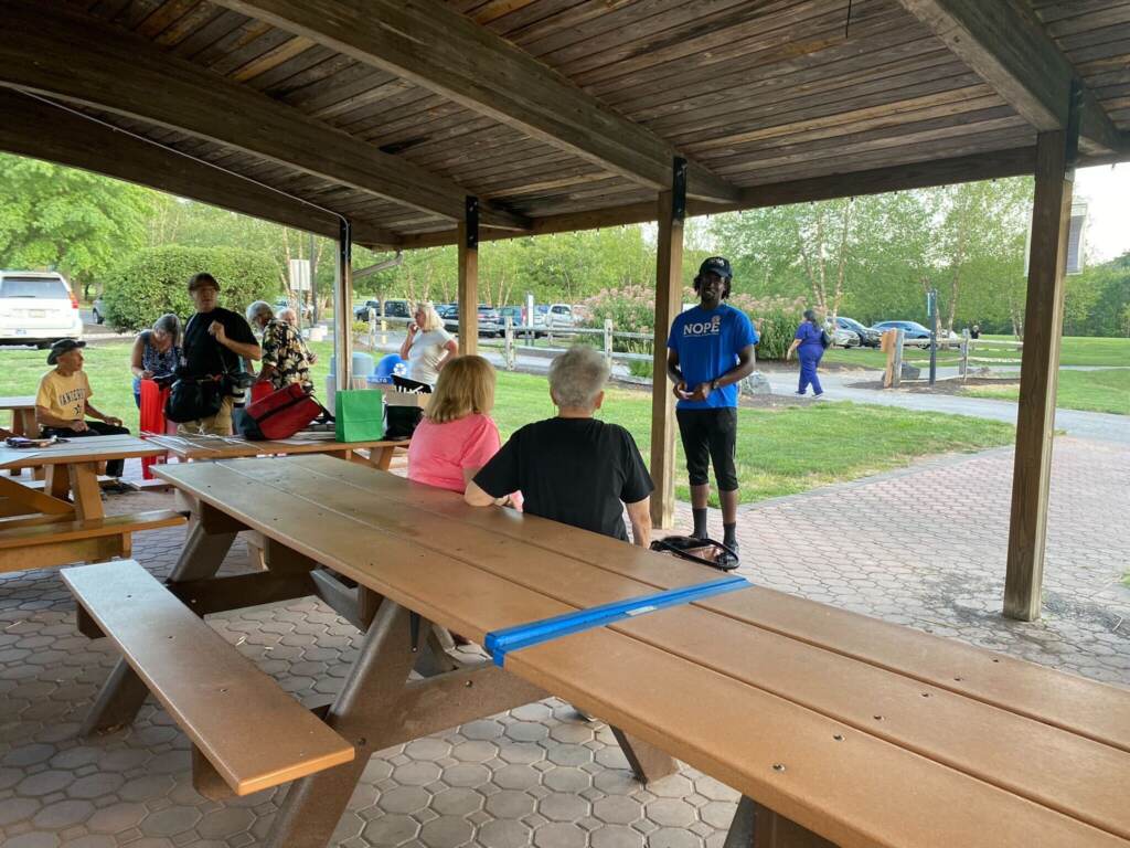 A man talks in front of a group of people seated at picnic tables in an outdoors area.