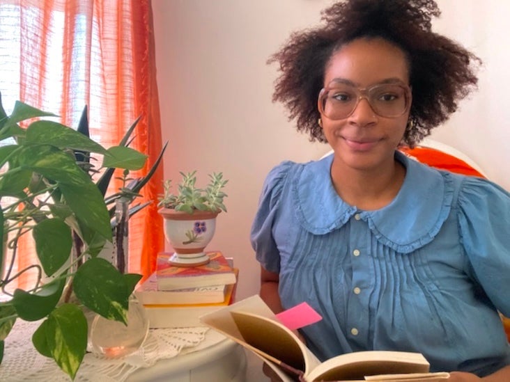 A woman smiles next to a desk, as she holds an open book.