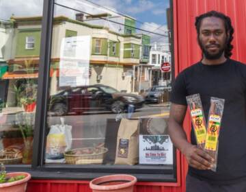 A man stands outside of a store, with the reflection of other stores on the street visible in the storefront window.