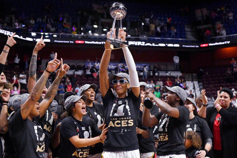 A basketball player holds up a trophy as her teammates surround her to celebrate.