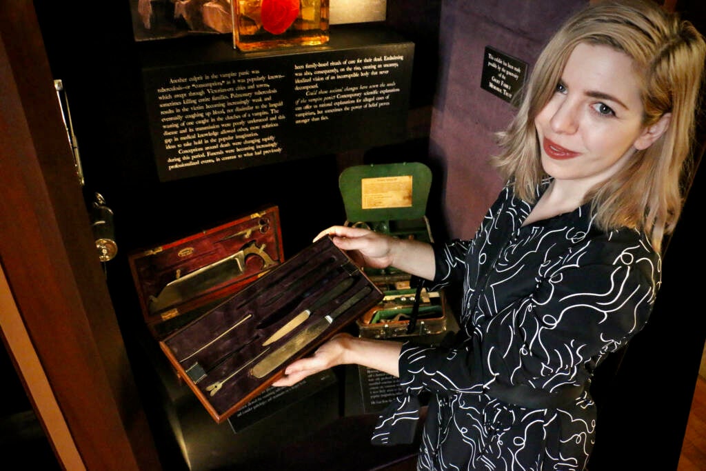 A woman displays a case of surgical instruments in front of an exhibit.