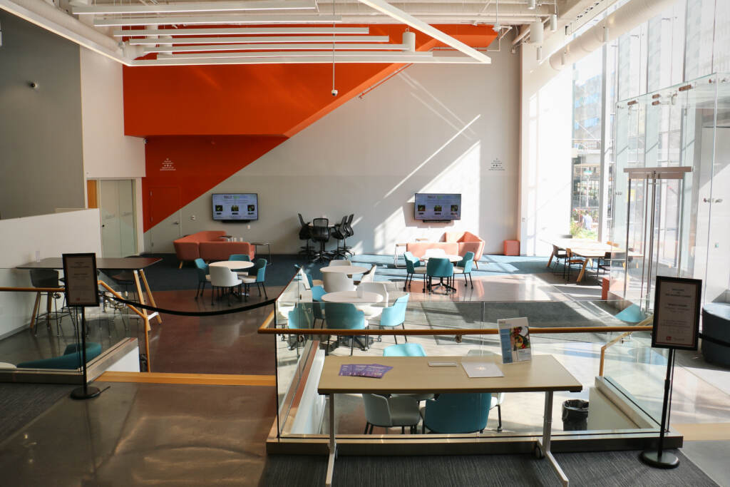 A view of a room, with tables, chairs, and light pouring in through tall front windows.