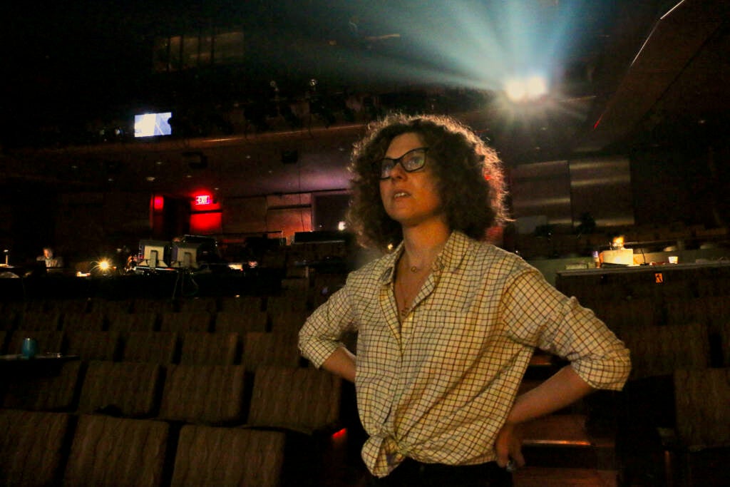 A person stands in a theater, a spotlight streaming behind her.