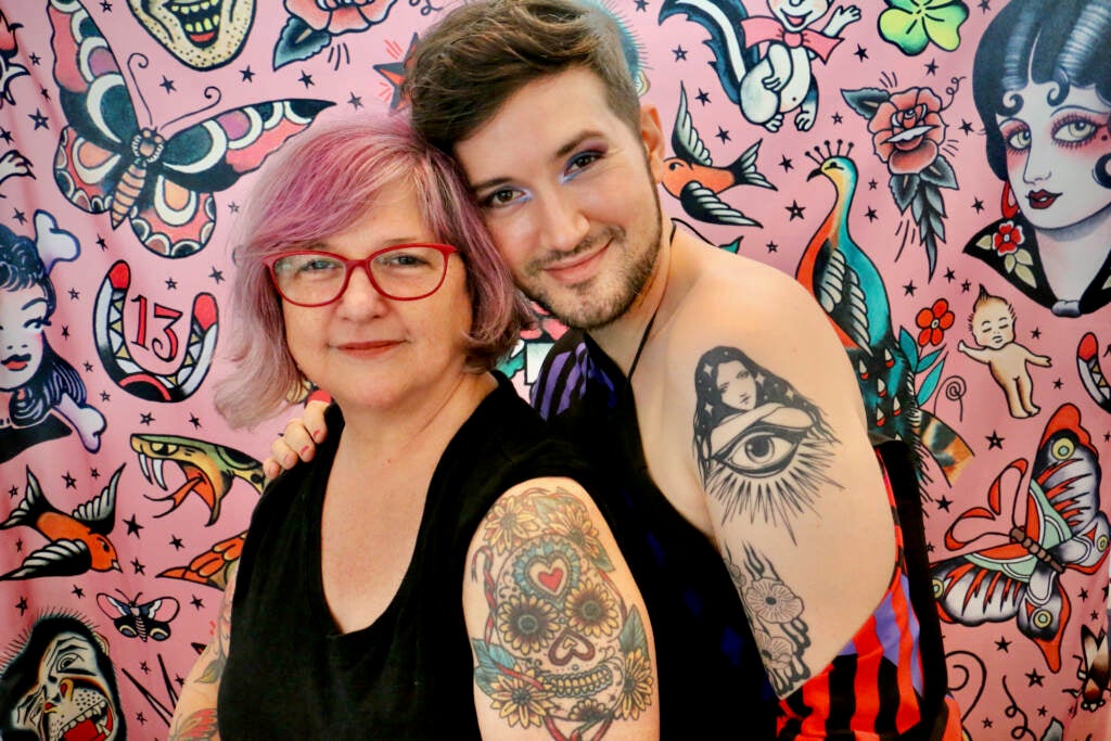 Two people face the camera, smiling, standing close, both with tattoos showing on their left arms.