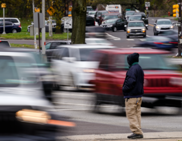 A silhouette of someone standing on a sidewalk is clear as traffic in the background going by is blurred.