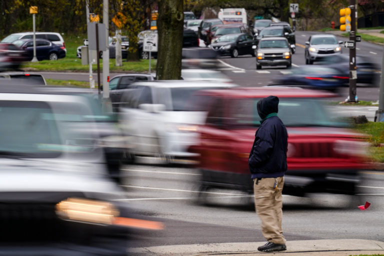 A silhouette of someone standing on a sidewalk is clear as traffic in the background going by is blurred.