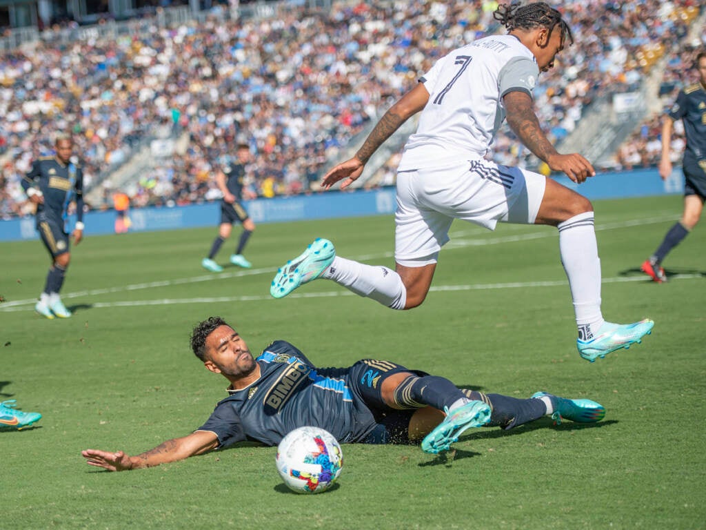 A soccer player is on the ground near the ball as a player on the opposing team jumps over him.