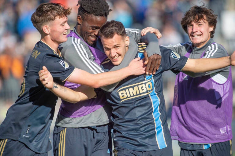 Soccer players surround and hug one soccer player who is smiling.