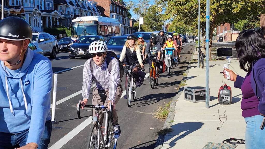 A line of bikers ride in a bike lane on a busy road.