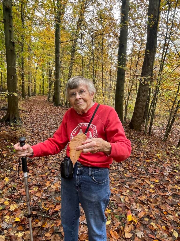 A woman holding a walking stick holds up a leaf with her other hand, as trees are visible behind her.