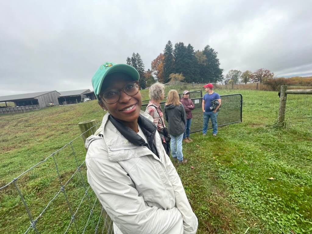 A woman smiles at the camera. Green fields and a group of people are talking behind her.