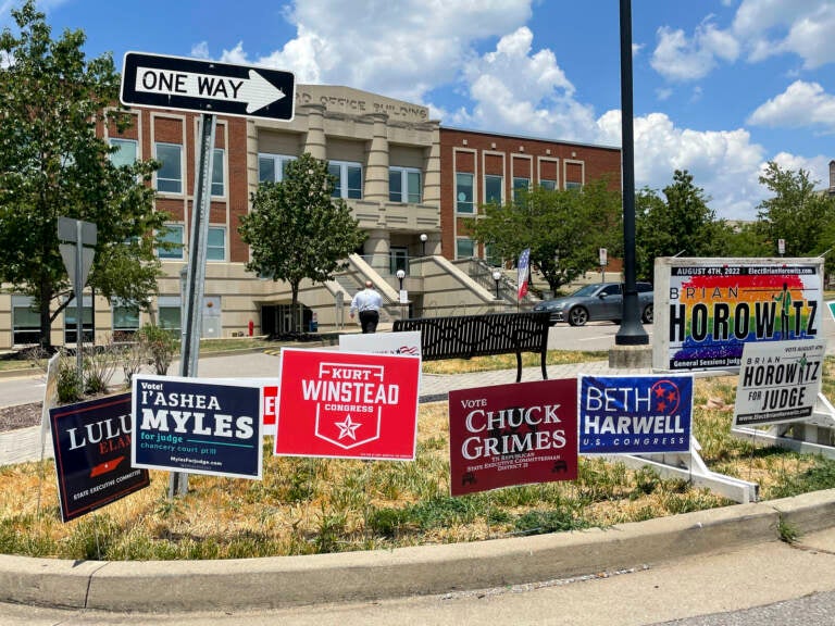 Political signs are visible stuck in a median strip of a road in front of a red brick building.