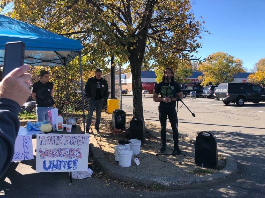 A person speaks at a microphone at a gathering in the parking lot of a Home Depot.