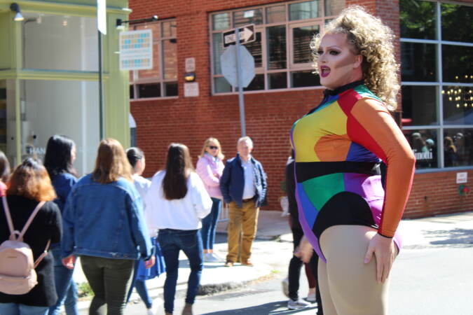 A performer dressed in a rainbow bodysuit sings as a crowd watches behind her.