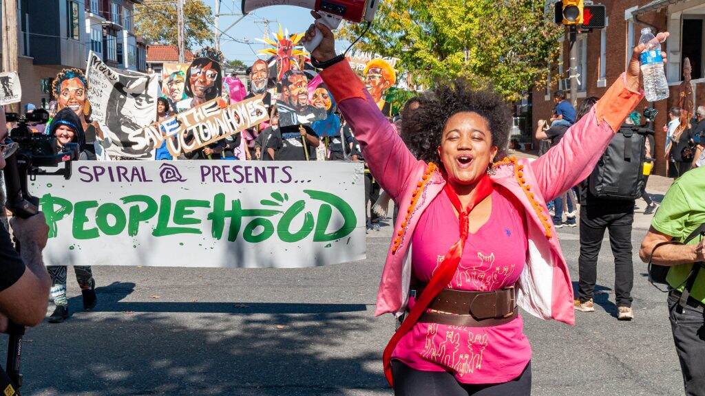 A person raises their arms as they walk forward. More people holding a sign that reads Peoplehood walk behind them.