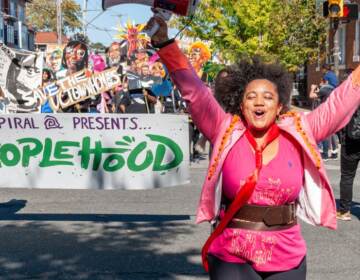 A person raises their arms as they walk forward. More people holding a sign that reads Peoplehood walk behind them.