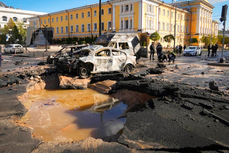 A busted up car sits near a crater in the street.