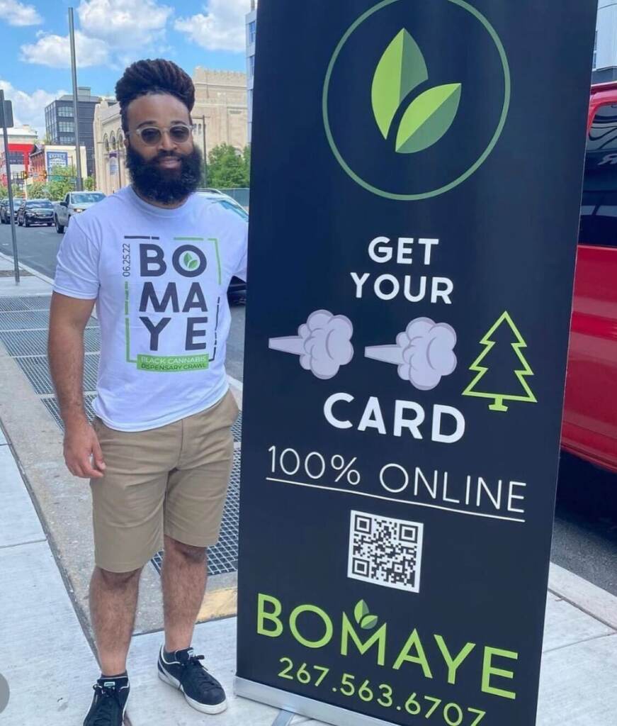 A man stands next to a sign that reads "Bomaye."