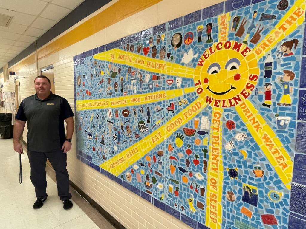 A man stands next to a mosaic of a sun that reads Welcome to Wellness.