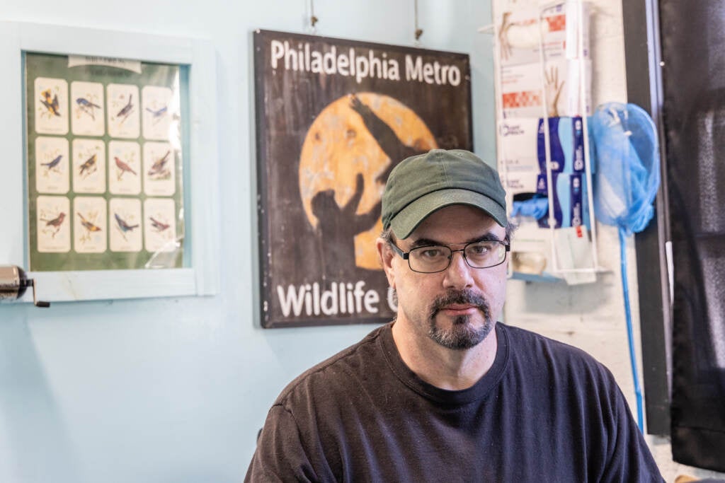 A man looks towards a camera. Behind him is a chart of birds and a sign that says Philadelphia Metro Wildlife.
