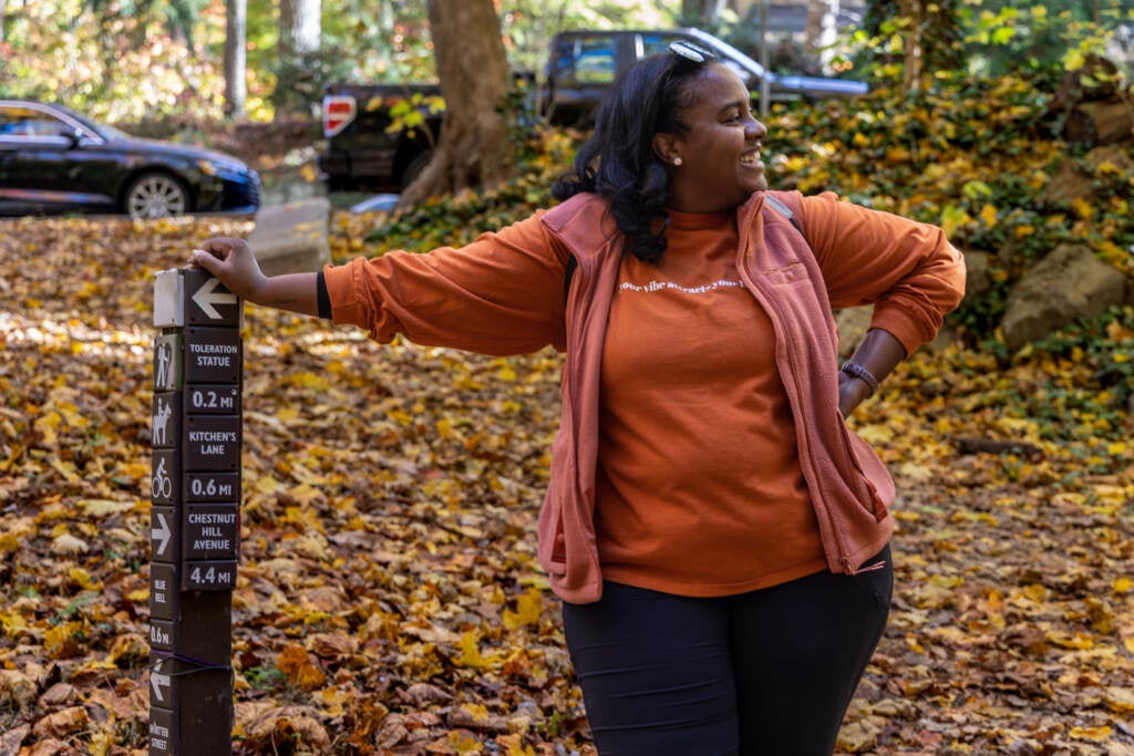 A woman leans on a trail post in the woods.