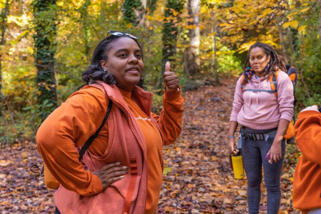 A woman poses, thumbs up while another person poses in the background.  Woods are visible behind them.