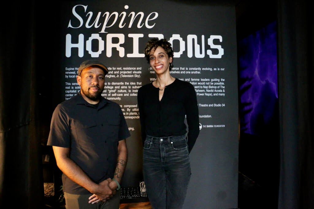Two people stand in front of a sign that reads, "Supine Horizons."