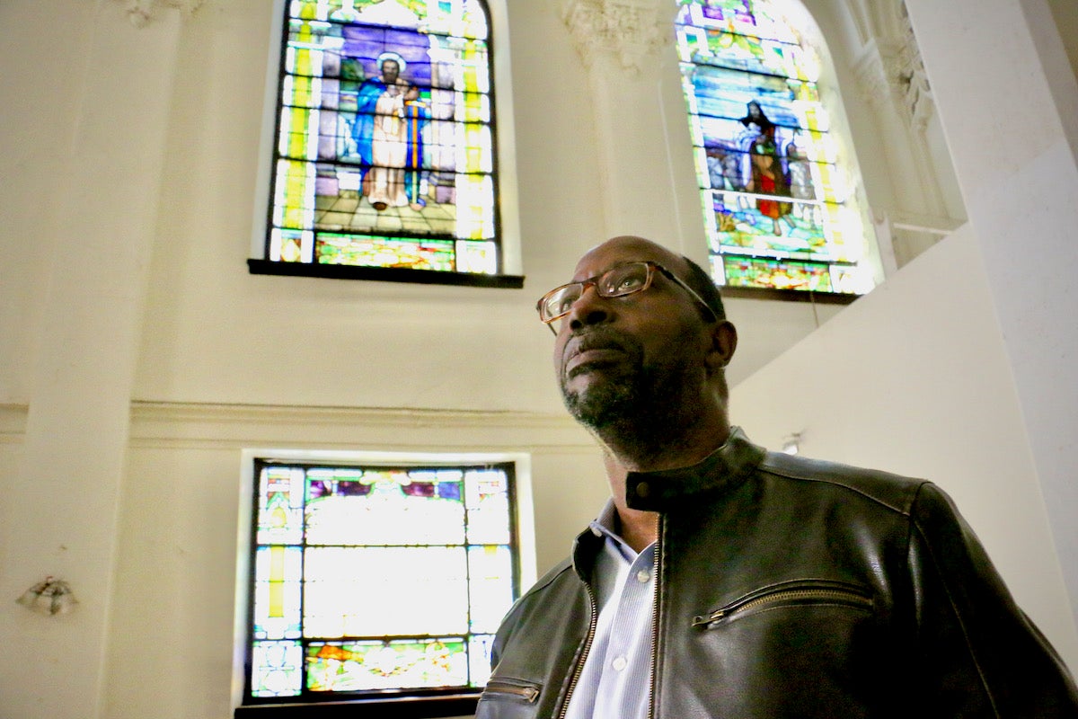 A man looks upward. Behind him are stained glass windows.