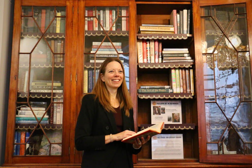 A person holds a book as she stands in front of a glass-paned bookshelf with an open door.