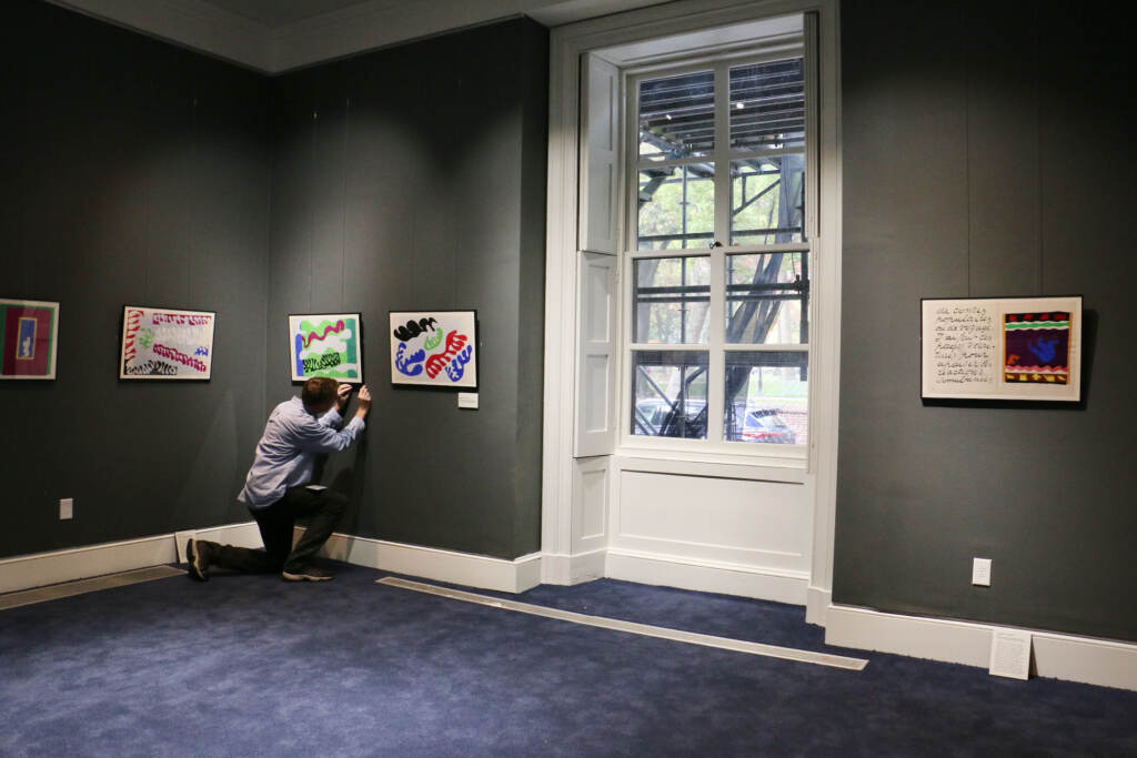 A person hangs paintings in a room with gray walls and a blue carpet.