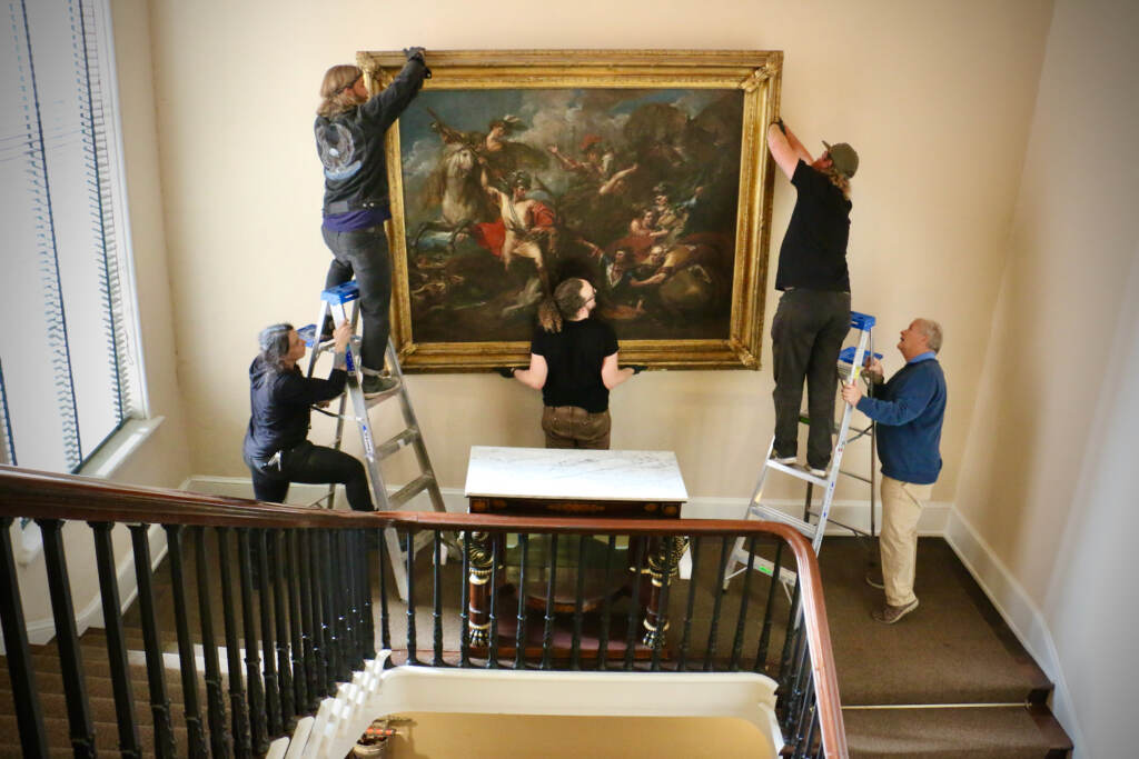 A group of people hang a painting on a staircase landing.