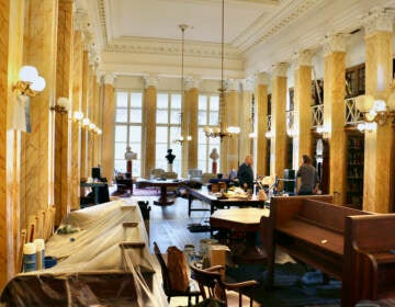 Workers make renovations in an ornate room with plastic draped over some of the desks.